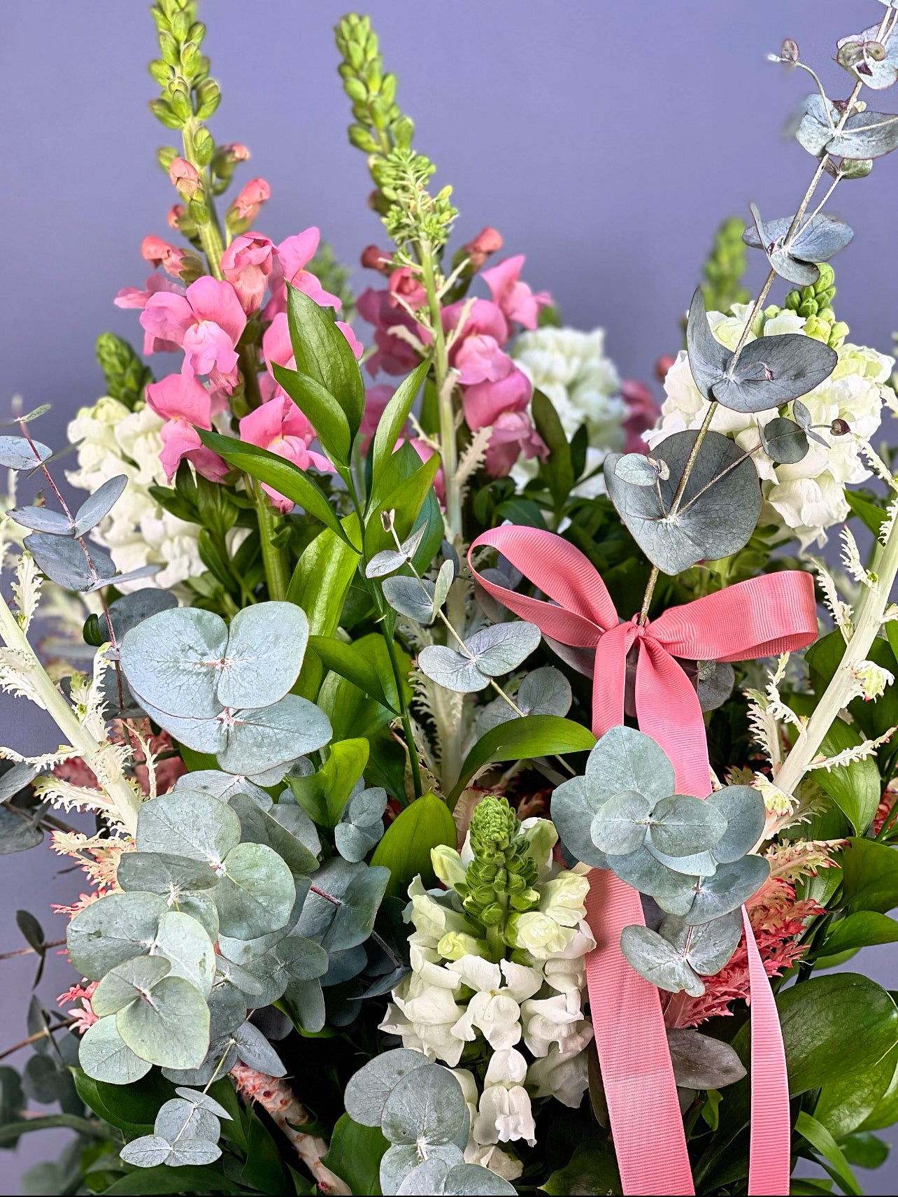 Yeşil cam vazonun modern tasarımıyla birleşen çiçek ve bitkilerin zarif düzenlemesi, aranjmanın sofistike atmosferini vurguluyor.