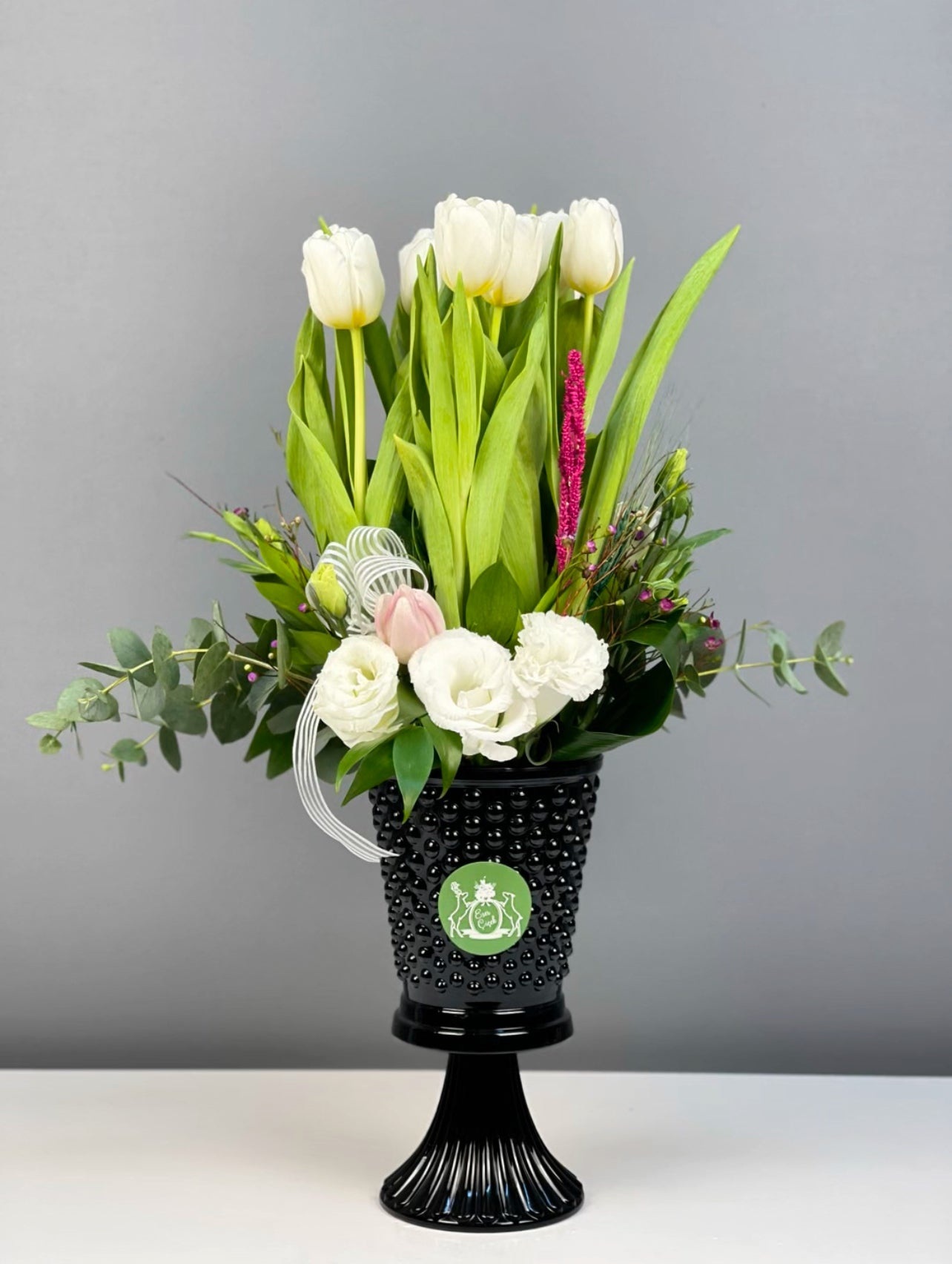 10 adet beyaz lale ve 3 adet beyaz lisyantusla oluşturulan Deluxe Cam Vazoda Ivory Harmony, 26 cm boy ve 14 cm ağız çapı siyah cam vazoda sergileniyor.