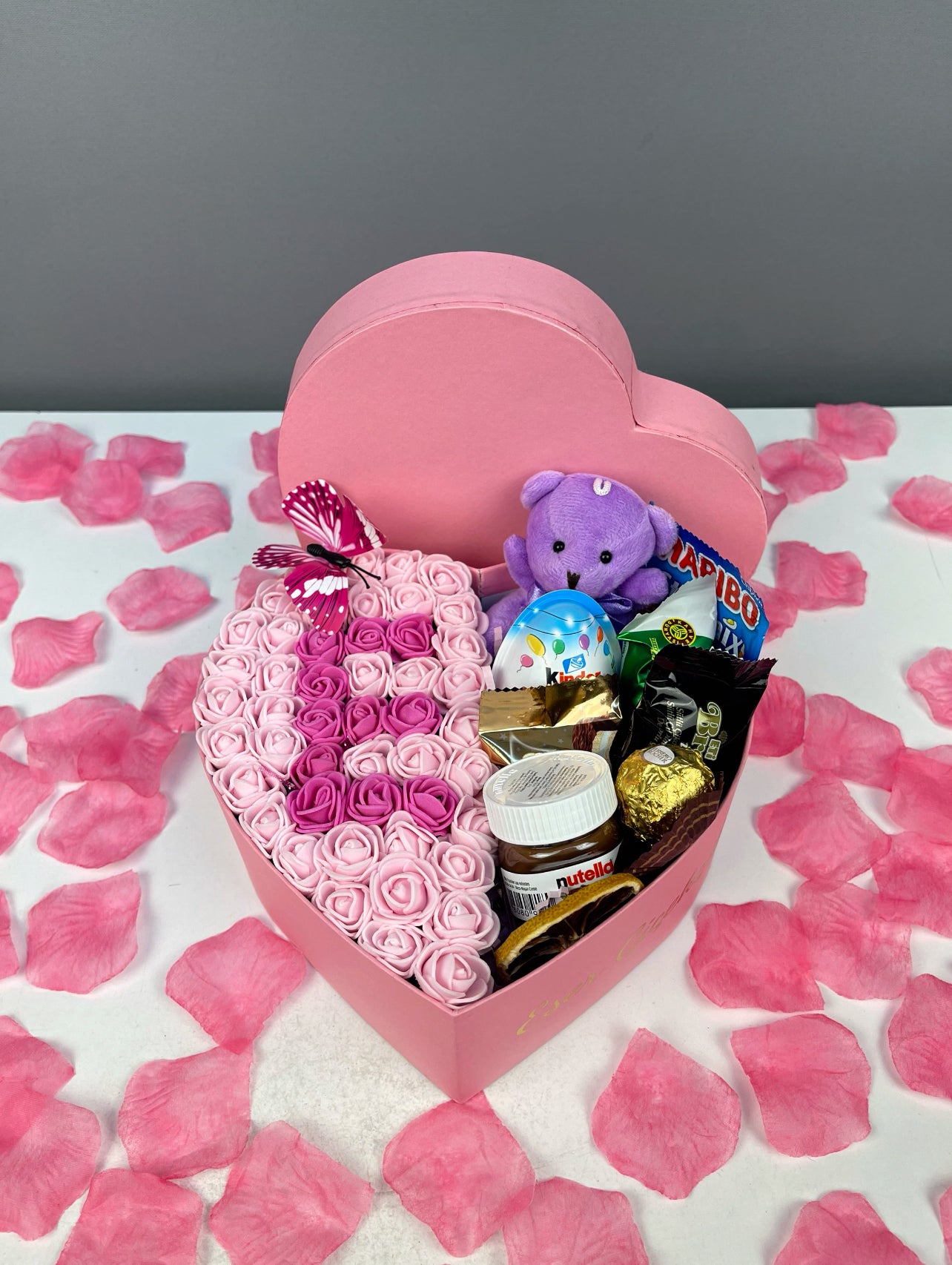 Çikolata ve peluş oyuncak bölümü ile fuşya güller üzerine pembe güllerden oluşturulan harf tasarımının üstten görünümü.